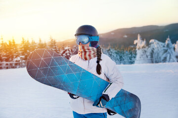 Portrait Of Woman On The Background Of Mountains and Sunset. Holding a Snowboard And Wearing Ski Glasses. Ski Goggles Of Snowboarder With The Reflection Of Snowed Mountains. Winter Sports