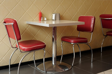 Classic diner scene featuring stylish red chairs around small table accented with salt, pepper, and ketchup. Diamond-patterned wall complements the retro vibe