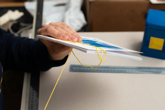 Artisan binding a book using traditional Japanese bookbinding techniques with yellow thread and white paper. The intricate stitching process, minimalist design, handmade craftsmanship.