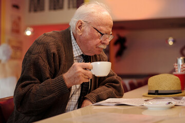 Senior man drinking coffee while reading newspaper in cozy cafe with relaxed atmosphere, wearing sweater and glasses