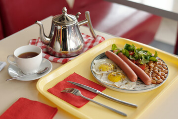 Breakfast tray featuring fried eggs, sausages, baked beans, and a fresh salad with a silver kettle and cup of tea on a red-checkered placemat and utensils
