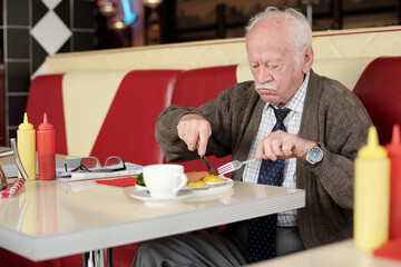 Elderly man enjoying meal in retro-style diner, with red and white seating accents and condiments on table. He is wearing a tie and sweater while focusing on his food