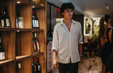 Stylish young man with curly hair browsing a wine collection at an elegant venue. The atmosphere is...