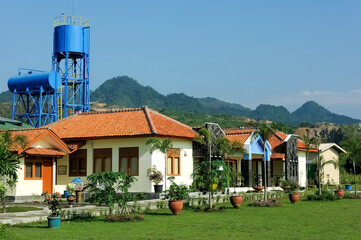 A serene villa surrounded by lush greenery, featuring a traditional yet modern architectural design with a sloping red-tiled roof. The villa is nestled amidst a garden landscape