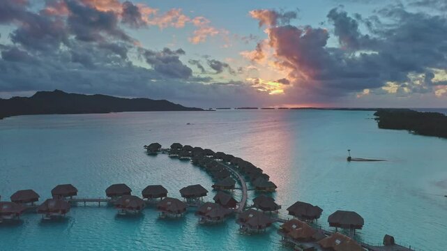 Luxury overwater bungalows resort on Bora Bora island. Wooden huts lined in turquoise ocean lagoon. Bright colorful clouds in sunset sky. Remote wild nature paradise, exotic summer travel. Aerial view