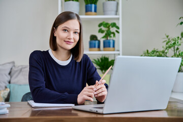 Portrait of young woman at her workplace at desk with laptop, looking at camera