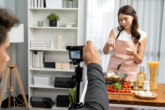 Cameraman recording to woman in chef influencer host cooking spaghetti with meat topped tomato sauce surrounded ingredients recipe, presenting special dish healthy food at modern studio. Postulate.