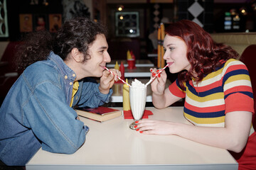 Young couple sharing milkshake at retro-styled diner with colorful decor while sitting across each other engaged in lively conversation