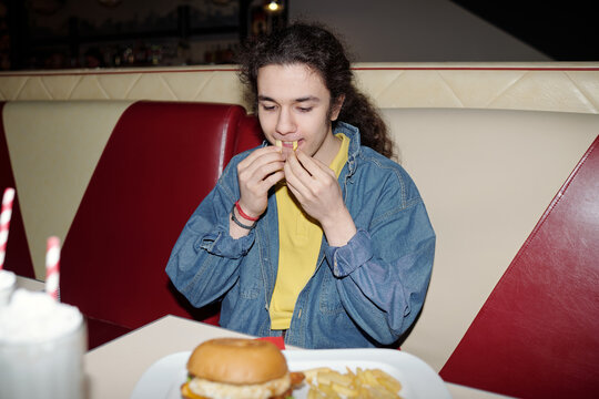 Portrait of teenager enjoying meal at retro-style diner booth, savoring french fries and ready to eat burger on plate without napkin or utensils present