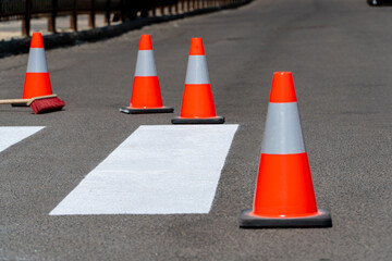 Bright orange traffic cones are placed along a newly marked crosswalk in an urban setting, directing pedestrians and ensuring safety in the area