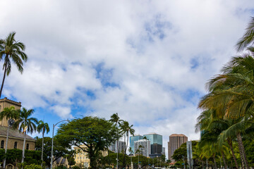 Hotels, condominiums and skyscrapers in the city skyline with lush green trees, cars driving on the street, blue sky and clouds in downtown Honolulu Hawaii USA