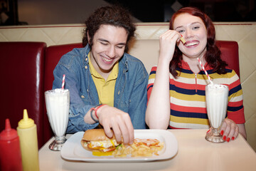 Couple sitting in cozy diner booth enjoying milkshakes, smiling and laughing, with burgers, fries, and condiments on table