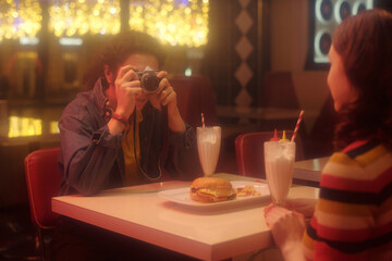 Person capturing food with camera at diner with milkshake and burger on table, another person...