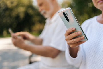 Close-up cropped shot portrait of beautiful elderly couple using smartphones sitting on park bench on sunny summer day, staying connected through technology amidst greenery. Concept of active aging