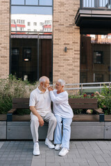 Full length vertical portrait of beautiful elderly couple enjoying happy moment sitting on city bench, embracing, warm talking and expressing love on summer day. Concept of retirement life.