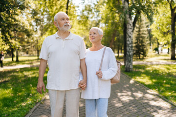 Obraz premium Portrait of loving senior couple enjoying leisurely stroll in green park, holding hands intertwined, sharing tender moment on sunny summer day. Active elderly people enjoying retirement together.