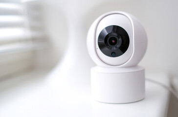 A white security camera with a black lens is mounted on a white surface. The camera is designed to capture images and record video footage