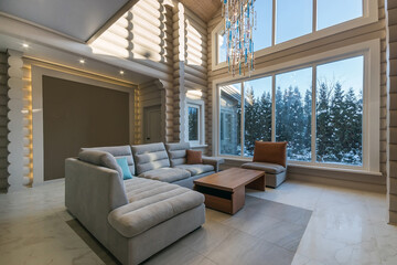 Living room in a luxurious wooden house with an exclusive chandelier on a high ceiling. .The panoramic window lets in the rays of the winter sun.