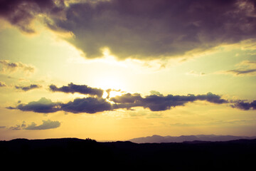 A breathtaking view of the sunset over Campos do Jordão, with the sun partially hidden behind clouds. The sky displays a gradient of warm colors, creating a serene atmosphere.