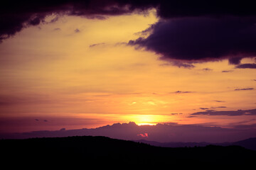 Beautiful sunset over rolling hills with a vibrant, colorful sky. The landscape features soft, dark silhouettes of the hills and a dramatic display of clouds.