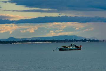 A rustic fishing boat anchored on calm waters with a scenic backdrop of distant mountains under a cloudy sky at sunset.