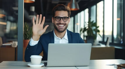 Fototapeta premium Portrait of a happy young businessman waving his hand and smiling while talking to the camera during a video conference call in the office, with a laptop in front of him. 