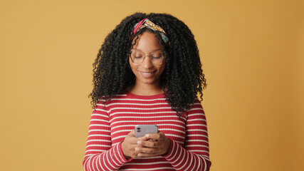 Young woman with curly hair wearing glasses wearing striped sweater, communicates with friends on social networks, mobile phone, on yellow background in the studio