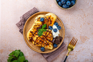 Homemade waffles with blueberries, chocolate sauce and cream cheese in a plate and a beige background top view