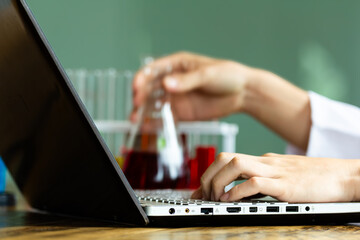 Close up male lab assistant chemist using laptop for data entry and in other hand holding test tube with red blood sample while sitting in central laboratory.
