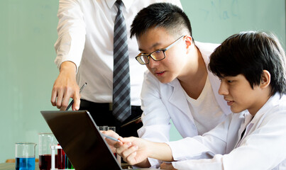 Young chemist student with teacher in school laboratory looking in laptop for new trial or research chemical. Working on university project together while studying in classroom.