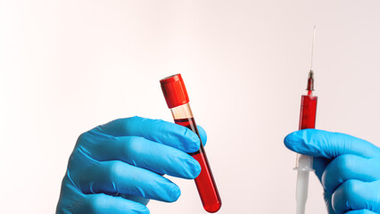 A person is holding a syringe and a blood sample. The syringe is red and the blood sample is also red. The person is wearing blue gloves