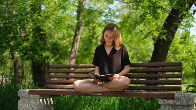 young woman student reading a book in the park on a bench on a sunny summer day