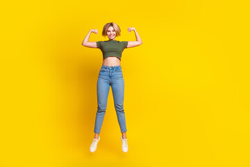 Full size photo of strong confident girl dressed khaki crop top jumping clenching fists show biceps isolated on yellow color background
