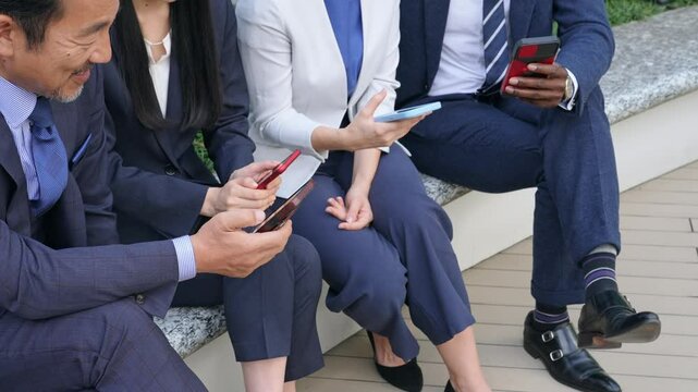 屋外ベンチに座ってスマホを見る多国籍ビジネスパーソンのグループ