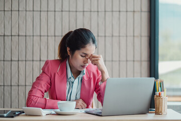 A businesswoman in a pink blazer appears stressed while working at her desk with a laptop and coffee in a modern office setting.