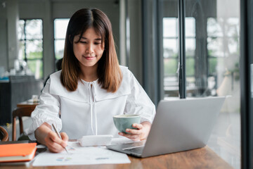 Obraz premium A young woman working remotely from home, using a laptop and holding a coffee cup, in a modern and bright home office environment.