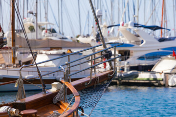 Obraz premium Bow of wooden sailboat in marina, surrounded by luxury yachts. Polished wood and chrome details, clear blue water and yachts in blurred background