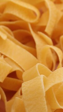 a close-up of raw, flat, wide noodles or pasta ribbons, rotation in circle. The pappardelle pasta is light golden in color with a slightly rough texture, Turning. Vertical video	 