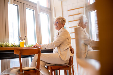 Joyful older woman works at a café, enjoying a refreshing drink and typing on her laptop in a...