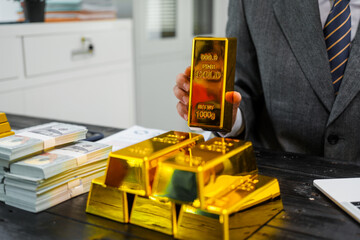 Businessman in suit sits at his desk, holding a shining gold bar. financial charts, the scene...