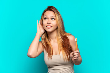 Teenager girl brushing teeth over isolated blue background listening to something by putting hand on the ear