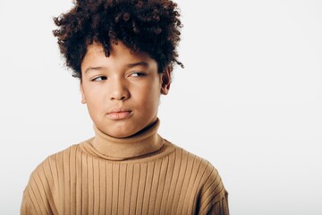 a charming portrait of a young african american boy with curly hair wearing a tan turtle neck sweater against a white background