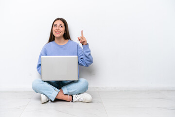 Young caucasian woman with laptop sitting on the floor isolated on white background pointing up and surprised