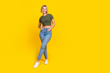Fototapeta premium Full size photo of adorable positive lovely girl dressed khaki t-shirt jeans holding arms in pockets isolated on yellow color background