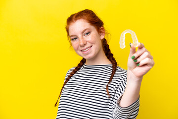 Young caucasian reddish woman holding invisible braces isolated on yellow background with happy expression