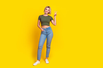 Full length photo of cheerful lovely girl dressed khaki t-shirt jeans arm in pocket show v-sign isolated on yellow color background