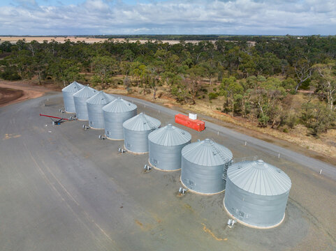 Aerial view of a line of grain storage bins in a clearing on the edge of a paddock