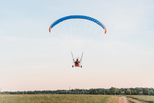 Paragliders on paraplane fly on red paraplane in sky above fields in countryside in summer at sunset