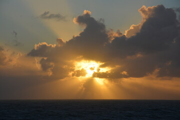 The sun sets behind dramatic clouds over the ocean, with golden rays breaking through the sky and...