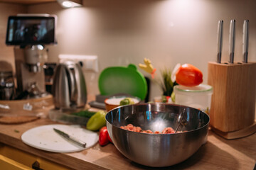 home kitchen countertop with ingredients for cooking, metal mixing bowl with minced meat, knives, vegetables, and kitchen appliances in the background